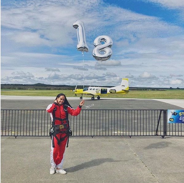 A woman in a red jumpsuit standing on a runway holding large silver number 18 balloons after receiving her birthday discount.